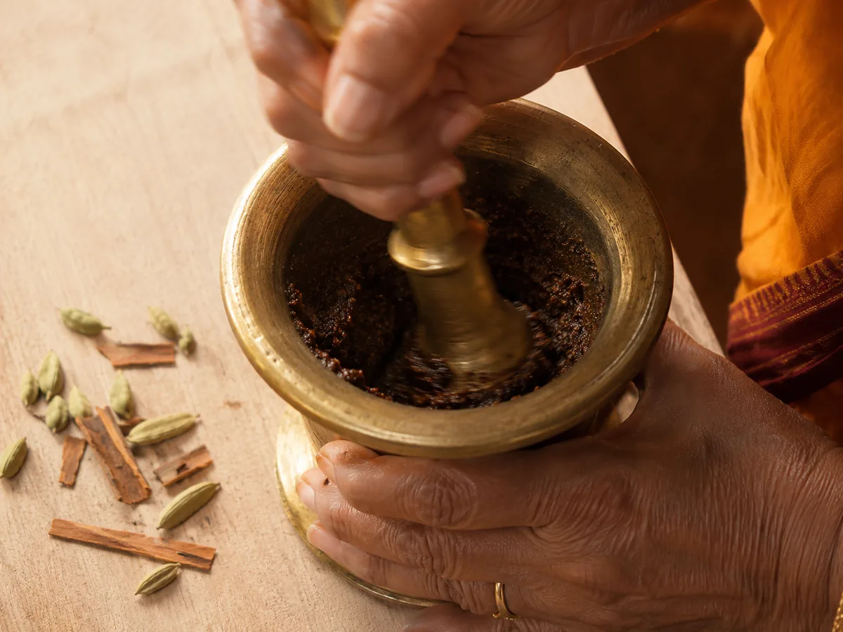 Two hands grinding masala in a brass mortar